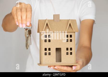 Hausschlüssel und ein kleines dekoratives Holzhaus in den Händen eines Mädchens auf weißem Hintergrund, Miete und Kauf von Immobilien, Haus und Wohnung Stockfoto