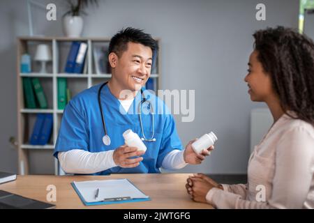 Glücklicher junger asiatischer Mann Arzt in Uniform berät schwarze Dame Patienten, bietet Gläser mit Pillen im Büro Stockfoto