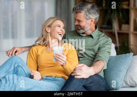 Portrait Eines Fröhlichen Paares Mittleren Alters, Das Mit Smartphone Auf Der Couch Ruht Stockfoto