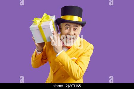 Fröhlicher, lustiger älterer Mann in hellen Kleidern, der eine Geschenkbox in den Händen auf purpurem Hintergrund hält. Stockfoto