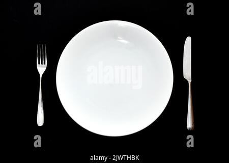 Messer und Gabel aus Edelstahl und leerer weißer Speiseteller auf schlichtem Hintergrund. Keine Personen. Stockfoto