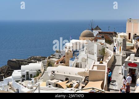 Oia, Santorini, Griechenland - Juni 2022: Menschen wandern durch die engen Straßen von Oia mit Gebäuden, die das Meer überblicken. Stockfoto