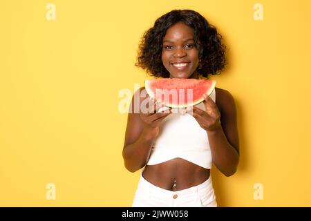 Foto von aufgeregt dunkle Haut Mädchen beißen natürliche Wassermelone Scheibe tragen weißes T-Shirt auf gelbem Hintergrund isoliert Stockfoto