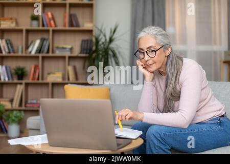Serious müde beschäftigt kaukasischen Senior grauhaarige Lehrerin in Brille arbeiten an Laptop im Wohnzimmer Interieur Stockfoto