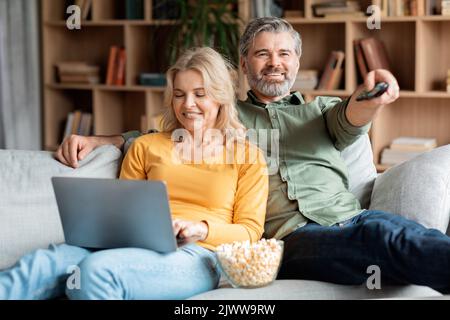 Reifes Paar, Das Sich Zu Hause Auf Der Couch Ausruht, Fernsieht Und Einen Laptop Benutzt Stockfoto