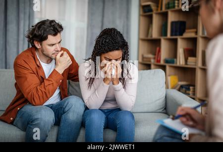 Verzweifelter trauriger junger europäischer Mann beruhigt die schwarz weinende Frau beim Treffen mit dem Psychologen im Büro Stockfoto