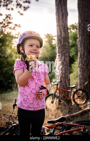 Ein kleines Mädchen mit einem Fahrradhelm, das vor dem Fahrrad steht, isst einen Apfel Stockfoto