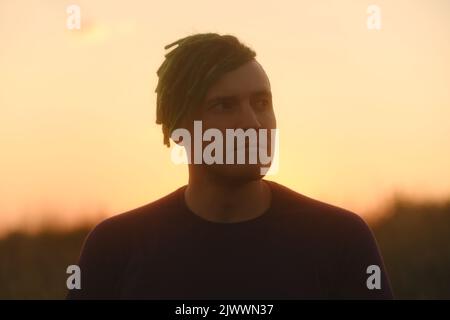 Junger, hübscher Mann mit Dreadlocks, der am Sonnenuntergang gegen den Himmel posiert Stockfoto