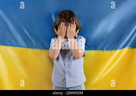 Hört auf, Kinder im Krieg zu töten. Ein kleiner verängstigte Junge weint, versteckt sein Gesicht mit Händen und posiert über der ukrainischen Flagge Stockfoto