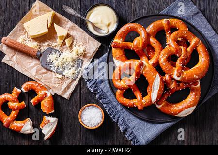 Weiche Brezeln in Form von Knoten gebacken und mit Salz auf schwarzem Teller auf dunklem Holztisch mit Käsesauce, geriebenem Käse und Reibe, hori bestreut Stockfoto