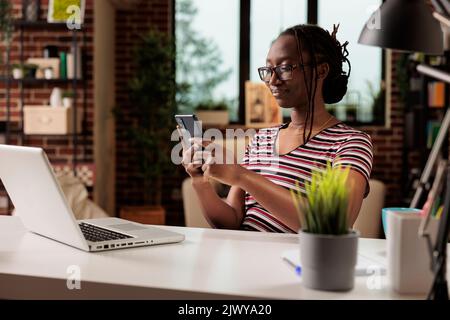 Lächelnde Frau Messaging auf dem Smartphone, chatten im sozialen Netzwerk am Schreibtisch des Heimbüros. Mitarbeiter mit Brille, der eine Pause von der Arbeit hat, Student, der auf dem Mobiltelefon im Internet surft Stockfoto