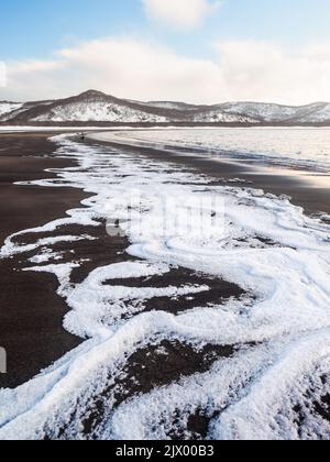 Meerwasser Schaum Wellen auf schwarzem Sand mit einem entfernten Berge Stockfoto