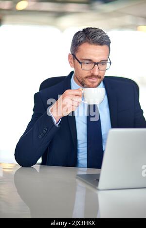 Lesen und Antworten auf seine E-Mails. Ein Geschäftsmann trinkt Kaffee und benutzt einen Laptop im Büro. Stockfoto