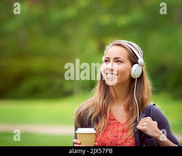 Machen Sie das Beste aus einem schönen Tag. Eine junge Frau, die Musik hört, während Sie unterwegs einen Kaffee trinken. Stockfoto