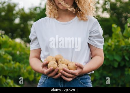 Zugeschnittenes Bild einer Frau in einem weißen T-Shirt mit frisch geernteten Kartoffeln. Arbeiter in der Landwirtschaft. Gesundes vegetarisches Essen, gesunder Lebensstil Stockfoto