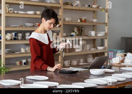 Junge beschäftigt fokussierte weibliche Unternehmerin mit Rechner, um Umsatz zu zählen, während der Arbeit in Keramik-Shop Stockfoto