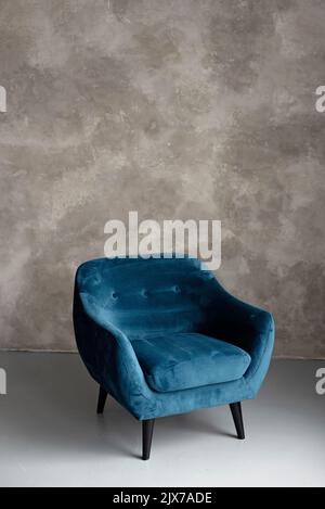 Klassischer blauer Samt-Art-Deco-Sessel mit Holzbeinen an einer grauen Wand. Vorderansicht. Weicher, selektiver Fokus. Stockfoto