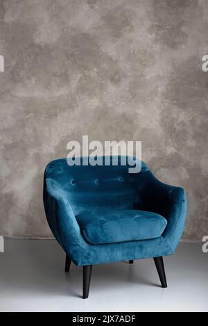 Klassischer blauer Samt-Art-Deco-Sessel mit Holzbeinen an einer grauen Wand. Vorderansicht. Weicher, selektiver Fokus. Stockfoto