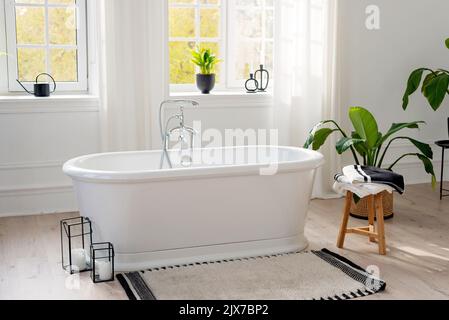 Stilvolle, moderne Badezimmereinrichtung. Horizontale Ansicht einer leeren freistehenden Badewanne auf einem Holzboden in einem hellen Raum vor dem Hintergrund eines großen Stockfoto