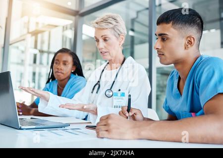 Doktor, der Medizinstudenten in einem Büro im Krankenhaus auf einem Laptop unterrichtet, während er Notizen schreibt. Erfahrener Professor mit Stethoskop-Ausbildung Stockfoto