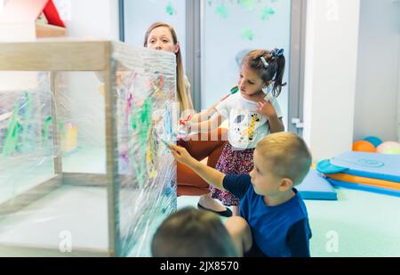 Frischhaltefolie Malerei für die Verbesserung der Kinder Phantasie und Gehirnentwicklung. Kleinkinder und ihre Lehrerin malen mit Bürsten, Rollen und Farben auf einer Frischhaltefolie, die um den hölzernen Regalständer gewickelt ist. Lustige Aktivität für Kinder feine und grobe Motorik im Kindergarten. Hochwertige Fotos Stockfoto