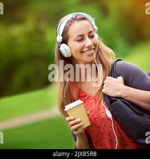 Ein Spaziergang durch den Park mit ihrem Kaffee und ihrer Musik. Eine junge Frau, die Musik hört, während sie unterwegs einen Kaffee trinken. Stockfoto