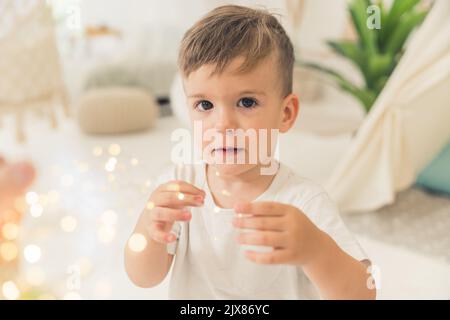 Nahaufnahme eines Innenportraits eines niedlichen kaukasischen, braunhaarigen und braunäugigen Vorschuljungen, der mit sicheren Dekorationsleuchten spielt und die Kamera anschaut. Verschwommenes Wohnzimmer im Hintergrund. Hochwertige Fotos Stockfoto