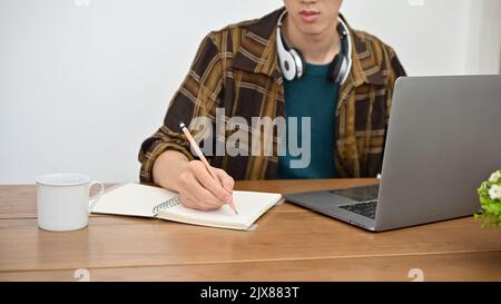Ein junger asiatischer Hipster-Student im Flanellhemd arbeitet an seinem Projekt, recherchiert Online-Informationen, benutzt Laptop und notiert sich h Stockfoto