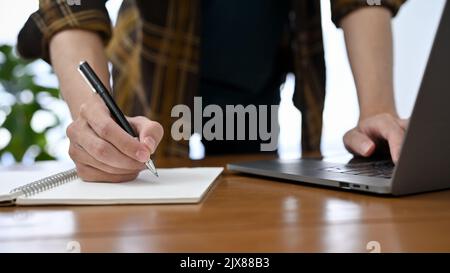 Ein professioneller Grafikdesigner, Freiberufler oder Büroangestellter in Flanellhemd, der an seinem Schreibtisch arbeitet, Notizen auf Spiralnotizbuch macht und mit Lapt arbeitet Stockfoto
