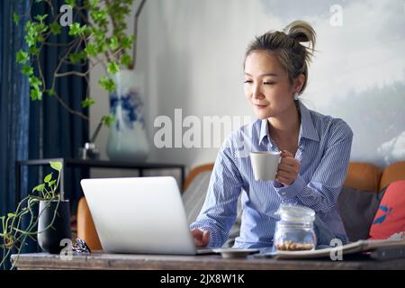 Reife professionelle asiatische Frau, die von zu Hause aus auf der Couch sitzt und Kaffee trinkt, während sie auf einen Laptop schaut Stockfoto