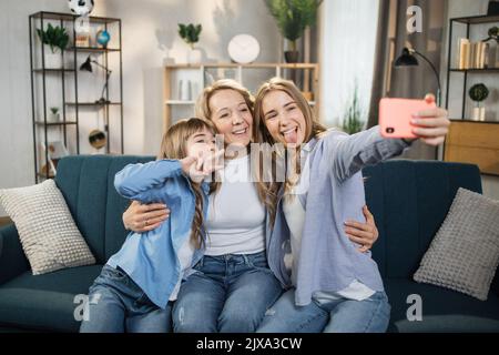 Foto-Porträt Nahaufnahme lächelnd zwei Töchter mit Mutter, die mit dem Smartphone Selfie macht, ihre Zungen zeigt, glückliche Mutter mit entzückenden Kindern, die Videoanruf machen oder Vlog aufnehmen Stockfoto