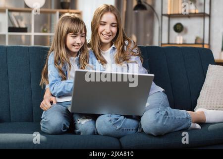 Kaukasische niedliche weibliche Kinder sitzen auf der Couch mit Laptop. Zwei Schwestern genießen Ferngespräche per Videoanruf, verbringen Zeit mit Internetkäufen, E-Commerce-Nutzern, sehen sich Cartoons oder Videofilme an. Stockfoto