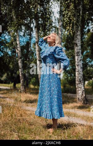 Eine junge Frau in einem langen Kleid steht an einem sonnigen Sommertag auf einer Lichtung. Stockfoto