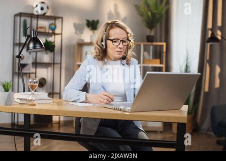 Portrait von schönen selbstbewusst mit blonden Haaren und auf Kopfhörern clevere Frau in legerer Kleidung, sitzen am Tisch vor dem Computer und Check Stockfoto