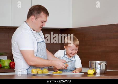 Konzept Nahrung und Ernährung. Aufnahme von zwei fröhlichen Papa und Sohn, die in der Küche posieren und Kartoffeln schälen, die zusammen vegetarisches Essen zum Abendessen kochen, mit identischen Schürzen Stockfoto