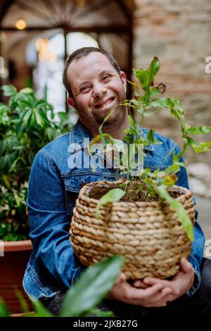 Junger Mann mit Down-Syndrom, der sich zu Hause um Pflanzen kümmert, lächelt und in die Kamera schaut. Stockfoto