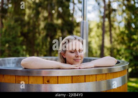 Junge Frau, die in der Holzbadewanne mit Kamin Holz verbrennt und Wasser im Hinterhof in den Bergen erwärmt. Stockfoto