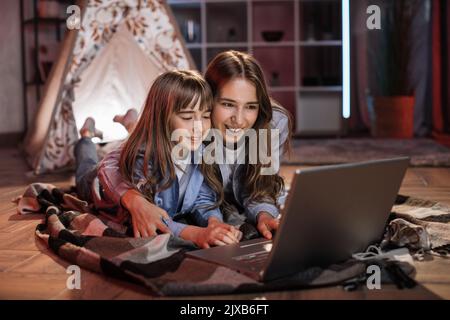 Schöne niedliche Schwestern oder Freunde, die auf dem Boden liegen, umarmen Wigwam mit einem Laptop. Glückliche Mädchen verbringen abends Zeit zusammen sehen Lieblingsfilm auf digitalem Gerät zu Hause. Stockfoto