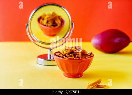 Mangochips in Schüssel mit Spiegel und roher Mango auf gelbem und orangenen Hintergrund Stockfoto