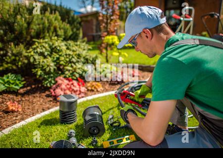 Professioneller Kaukasischer Gärtner mittleren Alters, der an der Bewässerungssystem-Installation im Garten seines Kunden arbeitet, indem er Schläuche schneidet, um sie zu entsprechen Stockfoto