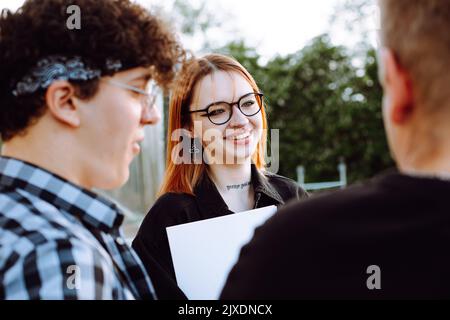 Nahaufnahme einer jungen, beschäftigten, verengten Gruppe von Menschen, zwei Männer und eine lächelte Frau in einer Brille machen Schreibarbeiten und reden im Freien Stockfoto