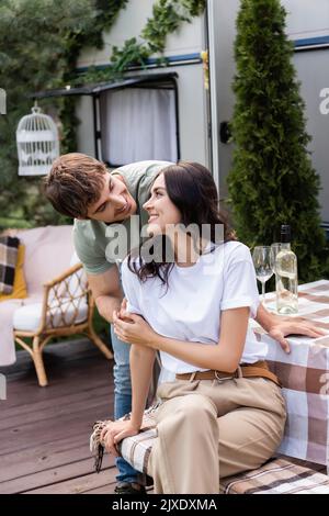 Lächelnder Mann umarmt Freundin in der Nähe von Wein auf der Terrasse des Wohnwagens, Stockbild Stockfoto