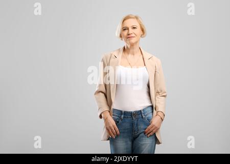 Selbstbewusste Frau im mittleren Alter, die mit Armen in den Taschen steht und auf grauen Hintergrund die Kamera anschaut Stockfoto