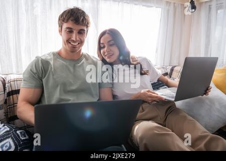 Positive Frau Blick auf Laptop in der Nähe verschwommen Freund auf dem Bett im Wohnmobil, Stockbild Stockfoto