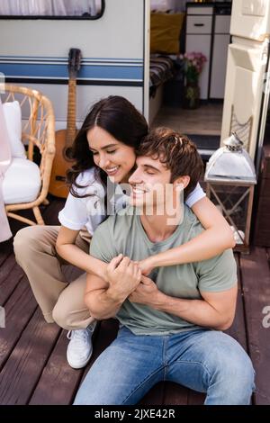 Junge Frau umarmt fröhlichen Freund in der Nähe Wohnmobil im Freien, Stockbild Stockfoto