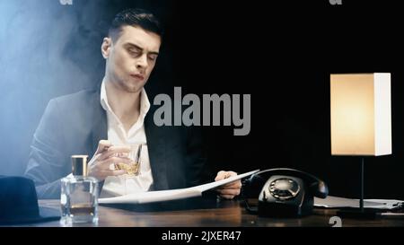 Geschäftsmann mit Whiskey und Notizbuch in der Nähe des Telefons und der Lampe auf schwarzem Hintergrund mit Rauch, Stockbild Stockfoto