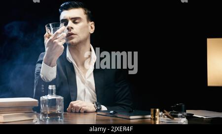 Geschäftsmann hält ein Glas Whiskey in der Nähe von Büchern und Zigarre in einem Aschenbecher auf Schwarz mit Rauch, Stockbild Stockfoto