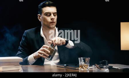 Ein Geschäftsmann hält eine Flasche Whiskey in der Nähe von Büchern und Aschenbecher auf einem schwarzen Tisch, Stockbild Stockfoto