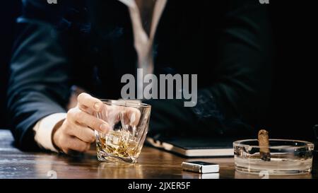 Beschnittene Ansicht eines verschwommenen Geschäftsmannes, der ein Glas Whiskey in der Nähe einer Zigarre in einem Aschenbecher hält, isoliert auf einem schwarzen Bild Stockfoto