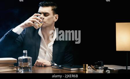 Geschäftsmann im Anzug trinkt Whiskey in der Nähe von Büchern und Aschenbecher auf dem Tisch isoliert auf schwarzem, Stockbild Stockfoto